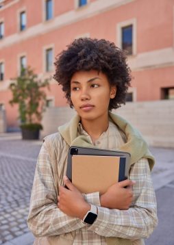 Vertical shot of beautiful curly haired woman carries notepad and tablet with app for students wears checkered shirt and vest walks oudoors against city building. Technology and lifestyle concept
