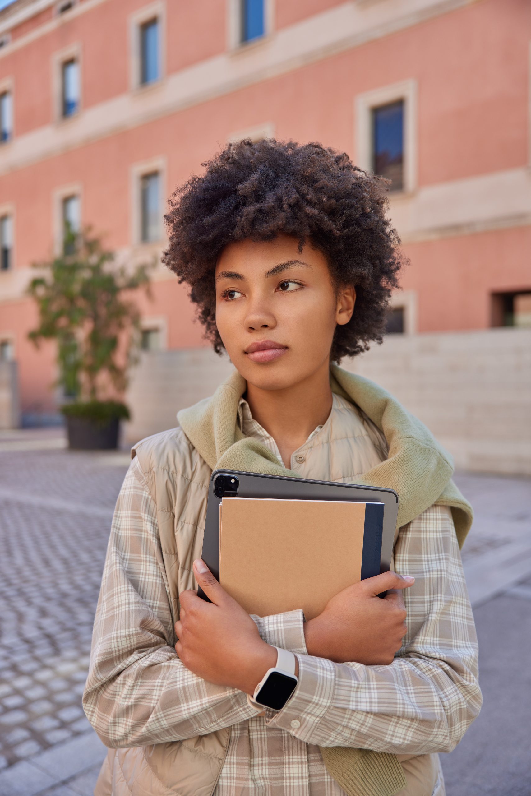 Vertical shot of beautiful curly haired woman carries notepad and tablet with app for students wears checkered shirt and vest walks oudoors against city building. Technology and lifestyle concept